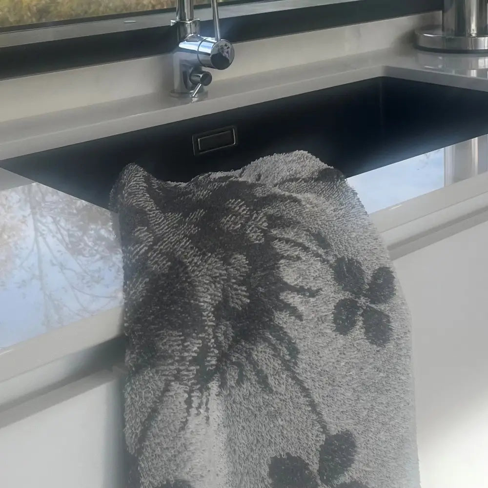 A plush gray käsipyyhe pellavafrotee with dark floral pattern hanging on a modern kitchen sink edge from jokipiin pellava
