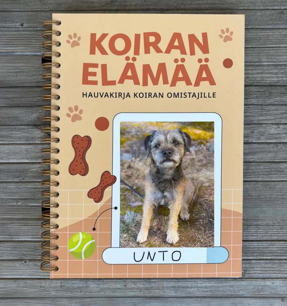 Beige kierrekiinnitteinen koiran omistajan päiväkirja oranssilla otsikolla, scruffy terrier-kuvalla sekä luu- ja tennispallokuvilla