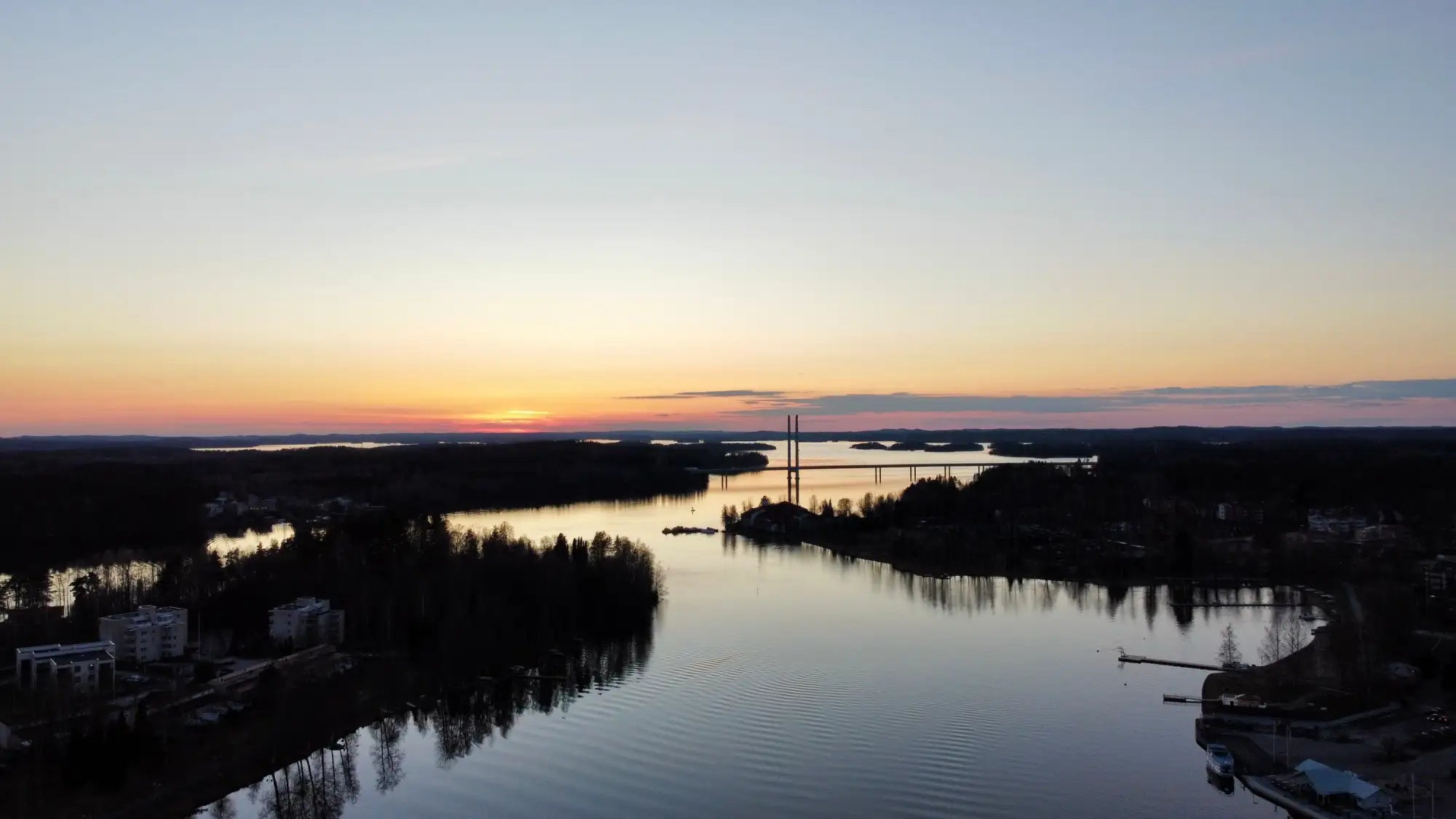 Raukea auringonlasku leveän joen yllä, taivas maalattu pehmeissä oranssin ja pinkin sävyissä siluetti-puiden ja kaukaisten rakennusten yläpuolella.