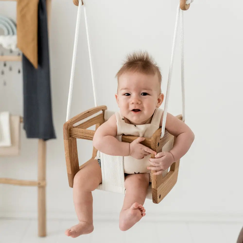 A baby sits in a toddler oak black dice wooden swing with white ropes and cream-colored fabric seat