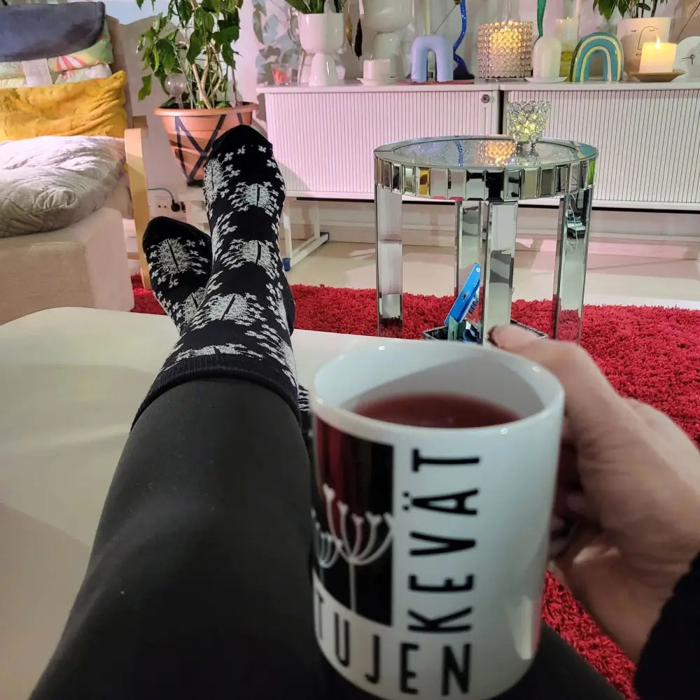 Valkoinen keramiikkamuki mustalla pystysuuntaisella tekstillä ja tyylitellyllä kukkakuvioinnilla, sisältää tumman punaisen juoman, pussin nimi tussukat tuhero puskapalovaara musta