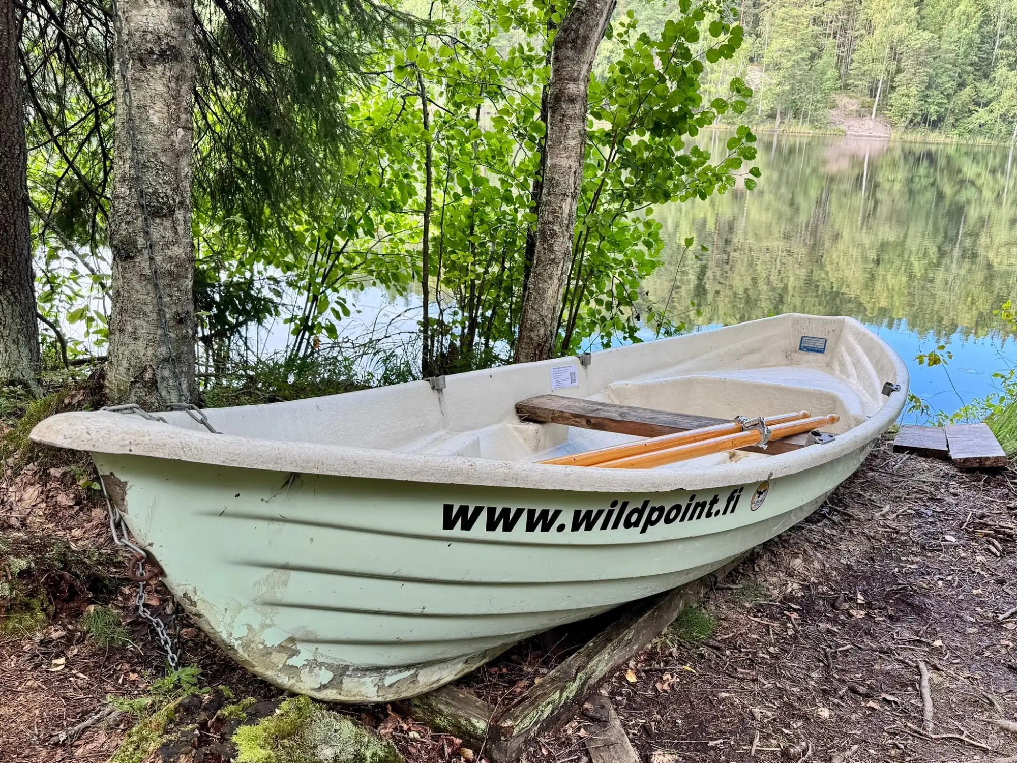 Vaalea vihreä, uritettu lasikuituinen soutuvene, puisilla aavereilla ja musta verkkosivustotarra sen kyljessä.