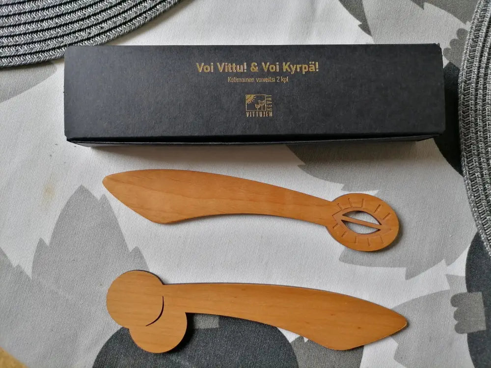 Kaksi vaaleanruskeaa puiset veitsenmuotoista koriste-esinettä kaiverretuilla kahvoilla voi vittu ja voi kyrpä lahjapakkauksessa